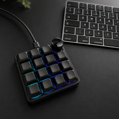 A compact 16-key mechanical keyboard pad with black keycaps and blue RGB backlighting, shown on a dark desk next to a computer keyboard.