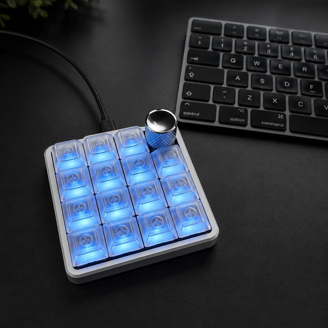 A silver color aluminum macropad controller with 16 glowing blue LED-lit buttons and a silver rotary knob, placed on a dark tabletop next to a keyboard.