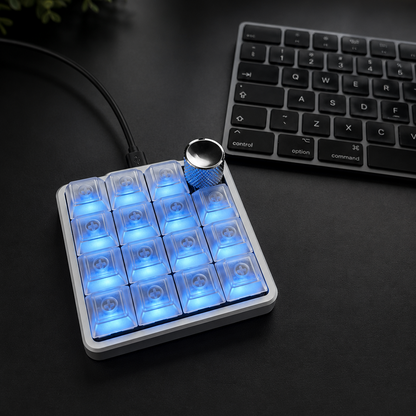 A silver color aluminum macropad controller with 16 glowing blue LED-lit buttons and a silver rotary knob, placed on a dark tabletop next to a keyboard.